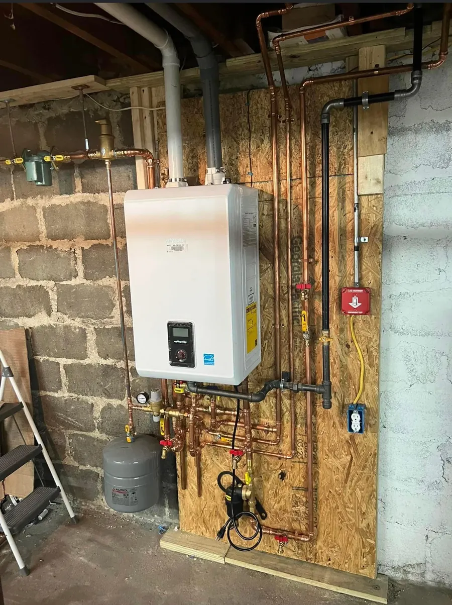 A white wall-hung boiler mounted on a plywood backboard, with copper and black gas piping and labelled valves below it, against a concrete block wall.